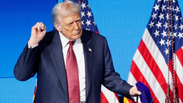 Donald Trump steht vor US-Flaggen, hält eine blaue Schachtel und gestikuliert.