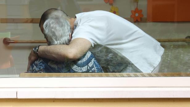Ein Mann in Weiß umarmt eine ältere Person mit grauen Haaren vor einem Fenster.