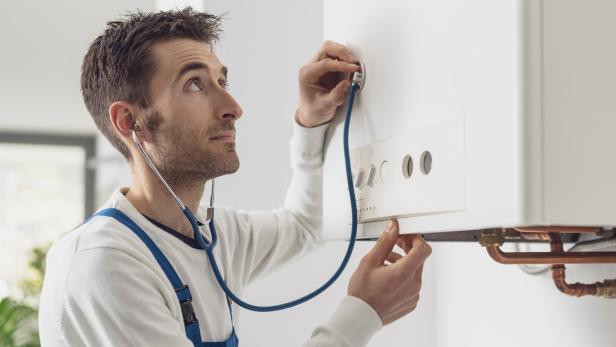 Handwerker mit blauem Stethoskop hört an weißem Heizkessel, trägt weißen Pullover und blaue Latzhose.
