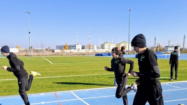Vier Personen laufen auf einer blauen Laufbahn im Freien bei sonnigem Wetter und tragen dunkle Sportkleidung.