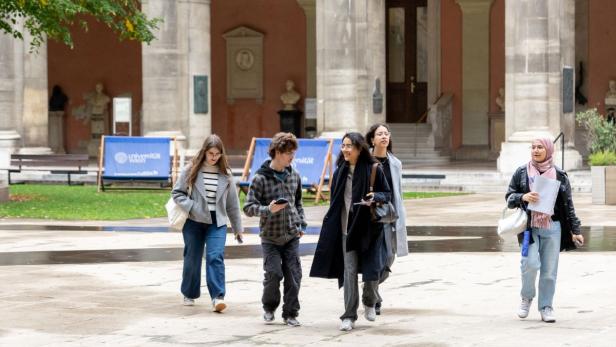 Gruppe von fünf Personen, die auf einem gepflasterten Platz vor einem historischen Gebäude mit Arkaden und Statuen (Arkadenhof der Uni Wien) gehen.