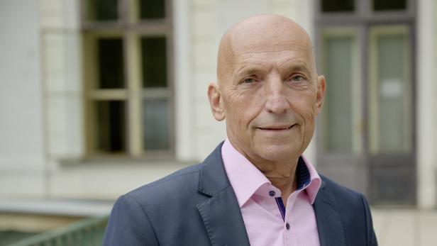 Ein älterer Mann mit Glatze trägt ein rosa Hemd und ein dunkles Sakko und steht vor einem Gebäude mit Fenstern.