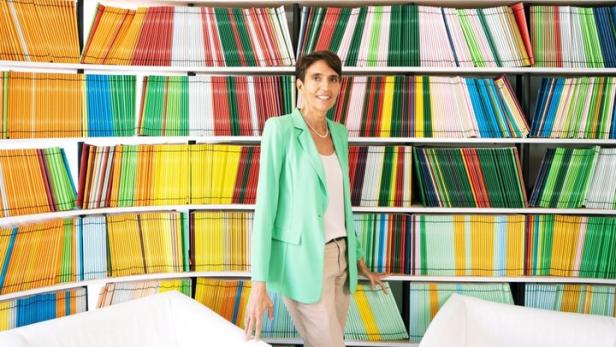 Eine Frau steht zwischen zwei weißen Sesseln vor einem Regal voller bunter, ordentlich sortierter Zeitschriften.