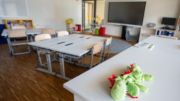 Ein grünes Stofftier in Drachenform liegt auf einem Tisch in einem modernen, leeren Klassenraum mit Tischen und Stühlen.