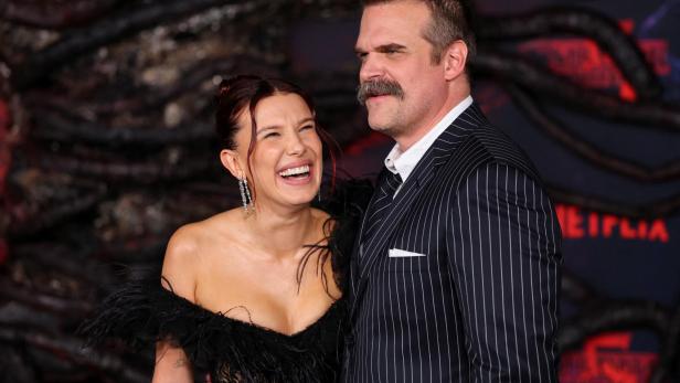 Millie Bobby Brown lacht neben David Harbour, der einen Nadelstreifenanzug trägt.