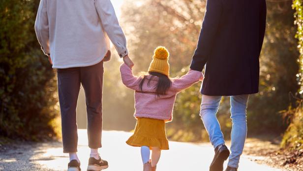 Ein Kind in gelber Mütze hält die Hände zweier Erwachsener und spaziert mit ihnen auf einem sonnigen Weg.
