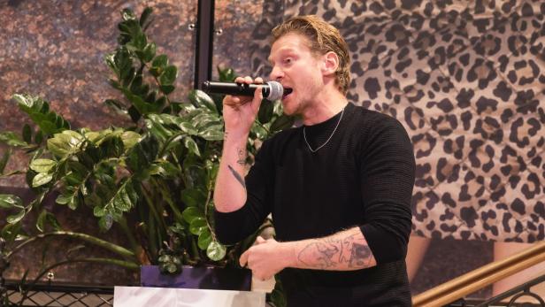 Ein Mann mit Tattoos singt mit einem Mikrofon, im Hintergrund sind grüne Pflanzen und ein Muster mit Leopardenflecken zu sehen.