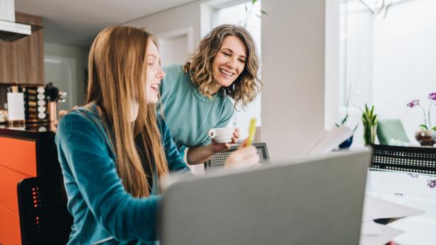 Mutter schaut ihrer Tochter über die Schulter während sie vor einem Laptop sitzt.
