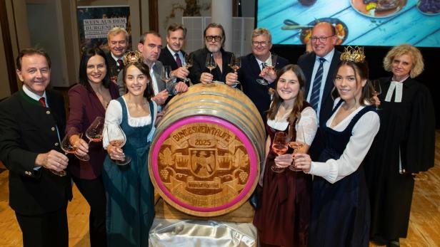 Mehrere Menschen stoßen feierlich mit Weingläsern um ein großes Weinfass mit der Aufschrift „Bundesweintaufe 2025“ an.