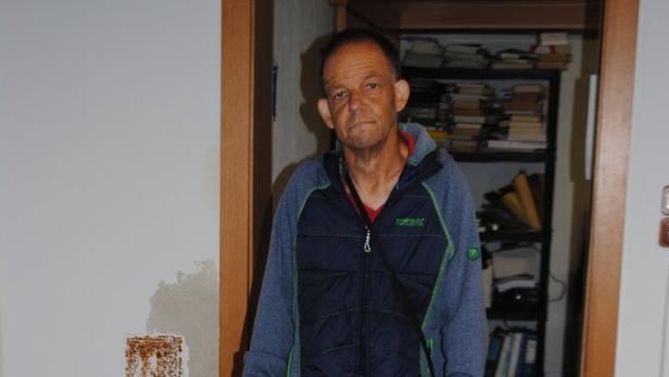 Ein Mann steht mit einem Rollator in einem Flur, im Hintergrund sind Regale mit Akten und Getränkeflaschen zu sehen.