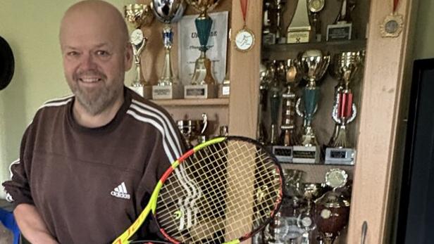 Ein Mann mit Beinprothese und Krücken hält einen Tennisschläger und steht vor einer Vitrine voller Pokale.