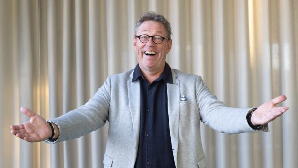 Ein Mann mit Brille und grauem Sakko steht lächelnd mit ausgebreiteten Armen vor einem hellen Vorhang.