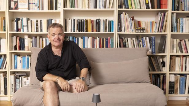 Cornelius Obonya sitzt lässig auf einer Couch vor seinem Bücherregal.