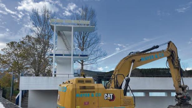 Ein gelber Bagger steht vor einem leeren Schwimmbecken mit Sprungturm, im Hintergrund herbstliche Bäume und blauer Himmel.