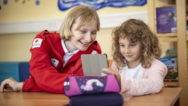 Frau in Rot-Kreuz-Jacke und Schulkind sitzen an Tisch