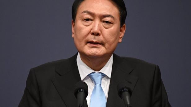 Ein Mann im Anzug mit hellblauer Krawatte spricht ernsthaft vor zwei Mikrofonen.