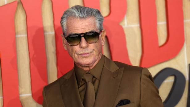 Pierce Brosnan mit Sonnenbrille und braunem Anzug vor rotem Hintergrund.