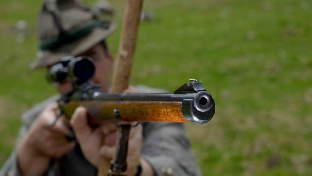 Eine Person mit Hut zielt mit einem Gewehr direkt nach vorne, der Lauf ist im Vordergrund scharf zu sehen.