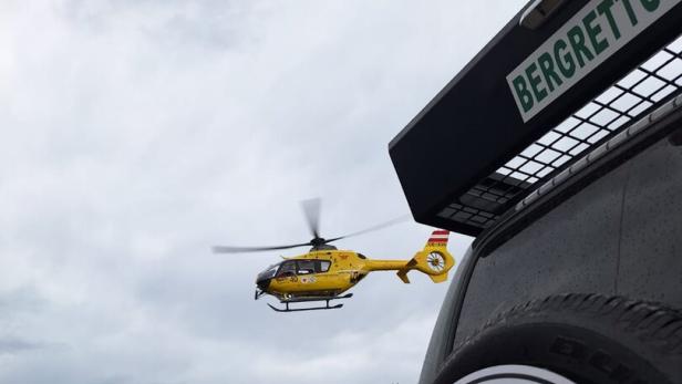 Ein gelber Rettungshubschrauber fliegt über eine Berglandschaft, im Vordergrund ein Fahrzeug der Bergrettung Österreich.