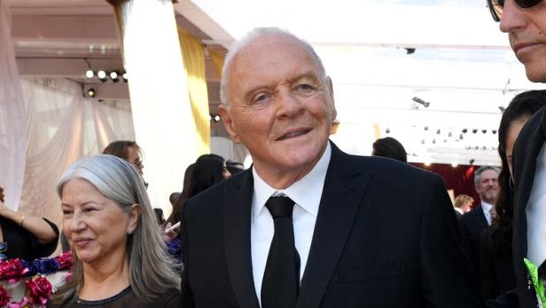 Ein älterer Mann im schwarzen Anzug und eine Frau im schwarzen Kleid gehen gemeinsam über einen roten Teppich.