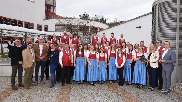 Eine große Gruppe von Menschen, viele in traditioneller Tracht, steht lächelnd im Freien vor einem modernen Gebäude.