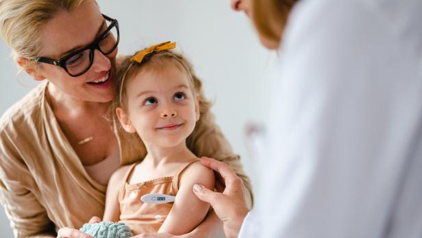Eine Mutter hält ihr Kind, während eine Ärztin mit einem Thermometer die Temperatur misst.