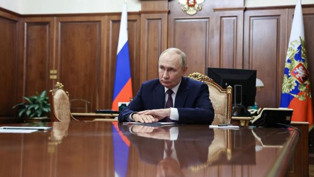 Putin sitzt an einem polierten Holztisch vor russischen Flaggen und dem Staatswappen.