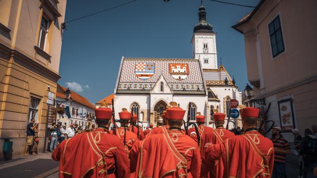Gruppe von Geistlichen in roten Uniformen mit goldenen Verzierungen vor der St.-Markus-Kirche in Zagreb mit farbigem Dach und Turm.
