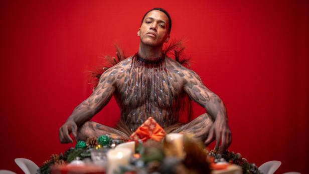 Eine Person mit bemaltem Körper und Federn sitzt auf einem festlich geschmückten Tisch vor rotem Hintergrund.