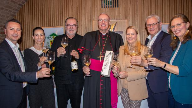 Sieben Personen stoßen mit Weißweingläsern an, eine Person trägt eine Bischofskleidung und hält eine Geschenkverpackung.