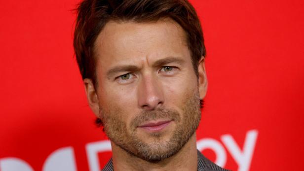 Glen Powell mit braunen Haaren und Bart schaut vor einem roten Hintergrund in die Kamera.