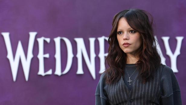 Jenna Ortega posiert vor dem Schriftzug "Wednesday" auf lila Hintergrund.