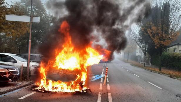 Ein Auto steht auf einer Straße in Flammen, dichter schwarzer Rauch steigt auf.