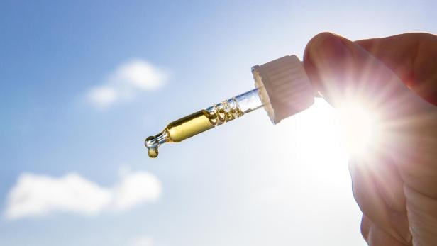 Eine Hand hält eine Pipette mit gelblicher Vitamind-D-Flüssigkeit vor blauem Himmel und Sonnenschein.