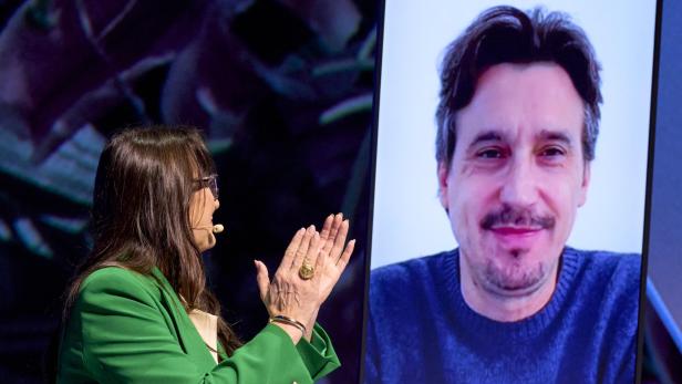 Eine Frau (Lilli Paasikivi) mit Mikrofon spricht mit einem Mann (Damiano Michieletto), der auf einem großen Bildschirm per Videoanruf zu sehen ist.