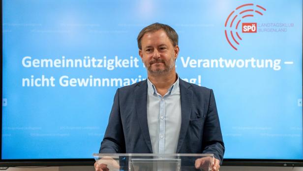 Ein Mann im Anzug steht an einem Rednerpult vor einem Bildschirm mit dem Text „Gemeinnützigkeit vor Verantwortung – nicht Gewinnmaximierung!“.