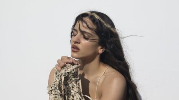 Rosalia mit langen dunklen Haaren trägt ein geflochtenes, cremefarbenes Kleid mit Fransen vor weißem Hintergrund.