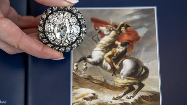 Eine Hand hält eine Diamantenbrosche vor einem Bild von Napoleon zu Pferde.