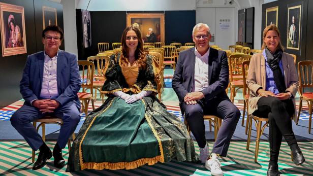 Vier Personen sitzen nebeneinander auf Stühlen, eine Frau trägt ein historisches Kleid, im Hintergrund sind Gemälde an den Wänden.