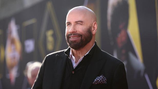 John Travolta lächelt mit Bart und Anzug vor einem unscharfen Hintergrund.