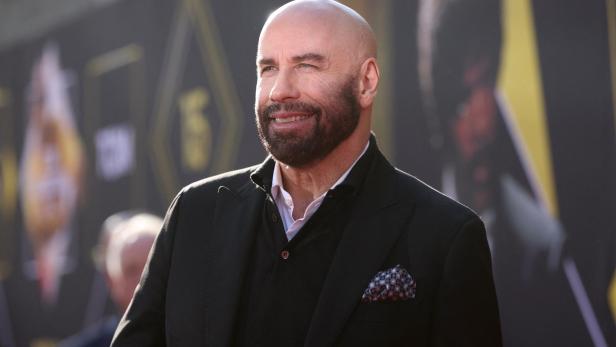 John Travolta lächelt mit Bart und Anzug vor einem unscharfen Hintergrund.
