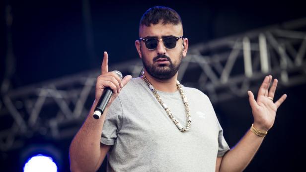 Rapper Haftbefehl mit Sonnenbrille, Goldkette und Mikrofon gestikuliert auf einer Bühne.