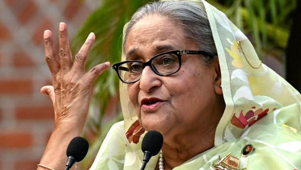 Sheikh Hasina mit Brille und Kopftuch gestikuliert vor Mikrofonen.