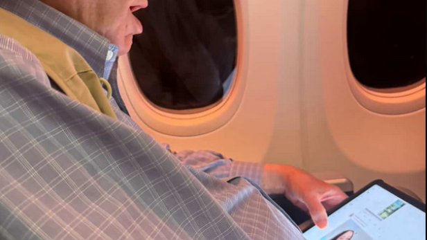 Brad Sherman sitzt im Flugzeug und betrachtet auf seinem Tablet ein Bild einer Frau in Unterwäsche.