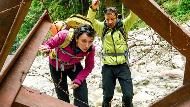 Felix Eitner an der Seite von Ursula Strauss mit Rucksäcken bücken sich vorsichtig unter Stacheldraht durch eine Metallkonstruktion im Gebirge.