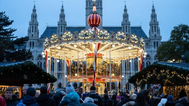 Ein beleuchtetes Karussell steht vor dem Wiener Rathaus, umgeben von Menschen und Marktständen.