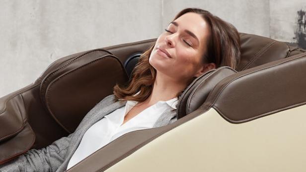 Frau mit braunen Haaren lehnt entspannt in einem braun-beigen Massagesessel, trägt grauen Blazer und weiße Bluse.