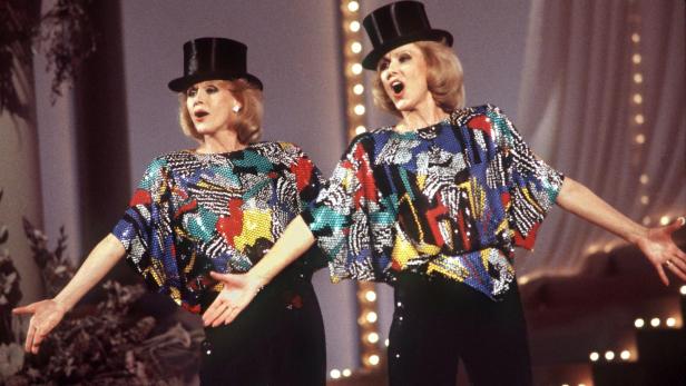 Zwei Frauen mit bunten Oberteilen und Zylindern singen und gestikulieren synchron auf einer Bühne.