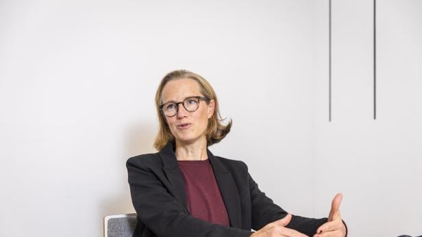Frau mit Brille und dunklem Blazer sitzt an einem Tisch und gestikuliert beim Sprechen.