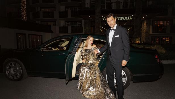 Ein Mann und eine Frau, die aus einem Auto steigen. Er trägt einen Smoking, sie ein goldenes Kleid. 